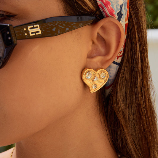 Gold heart-shaped stud earrings with embedded crystals, worn by a model with sunglasses and patterned headband – Orlaya Jewellery