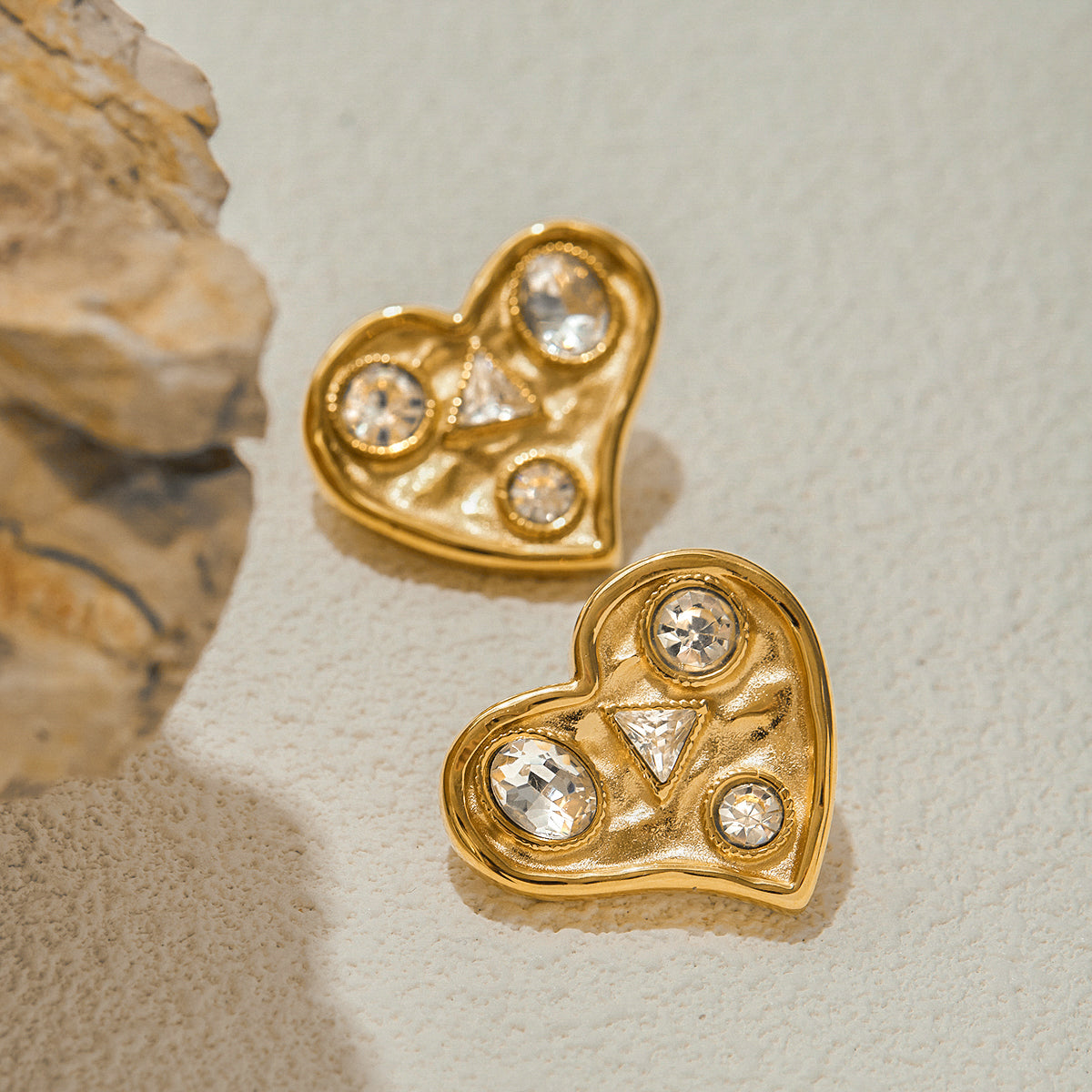 Gold heart-shaped earrings with embedded crystals on a textured surface