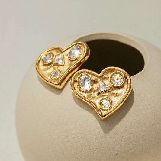 Gold heart-shaped stud earrings with embedded crystals, displayed on a beige ceramic surface – Orlaya Jewellery