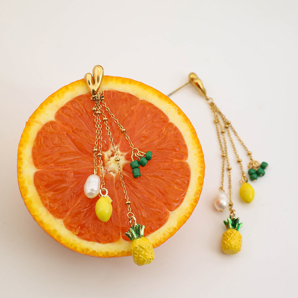 Close-up of gold pineapple and pearl dangle earrings with green bead accents on orange background – beach holiday earrings – Tropic Pop by Orlaya Jewellery