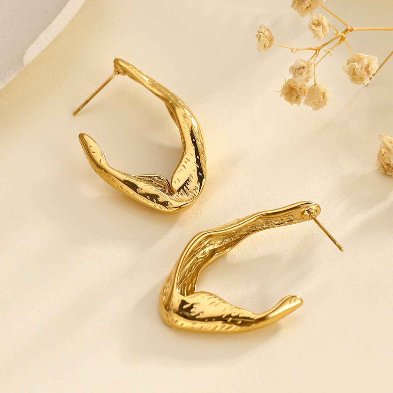 Pair of Zephyr gold sculptural hoop earrings styled with small flowers in the background